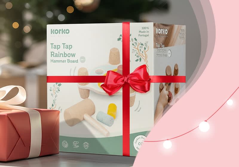Eine Geschenkbox mit rotem Band enthält ein Spielzeug namens „Tap Tap Rainbow Hammer Board“ von Korko, 100% Made in Portugal, auf einer rosa Fläche mit Lichterkette im Hintergrund