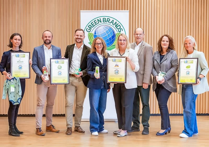 Eine Gruppe von sieben Personen steht vor einem Banner mit dem Logo von Green Brands, einige halten Zertifikate und Produkte in den Händen