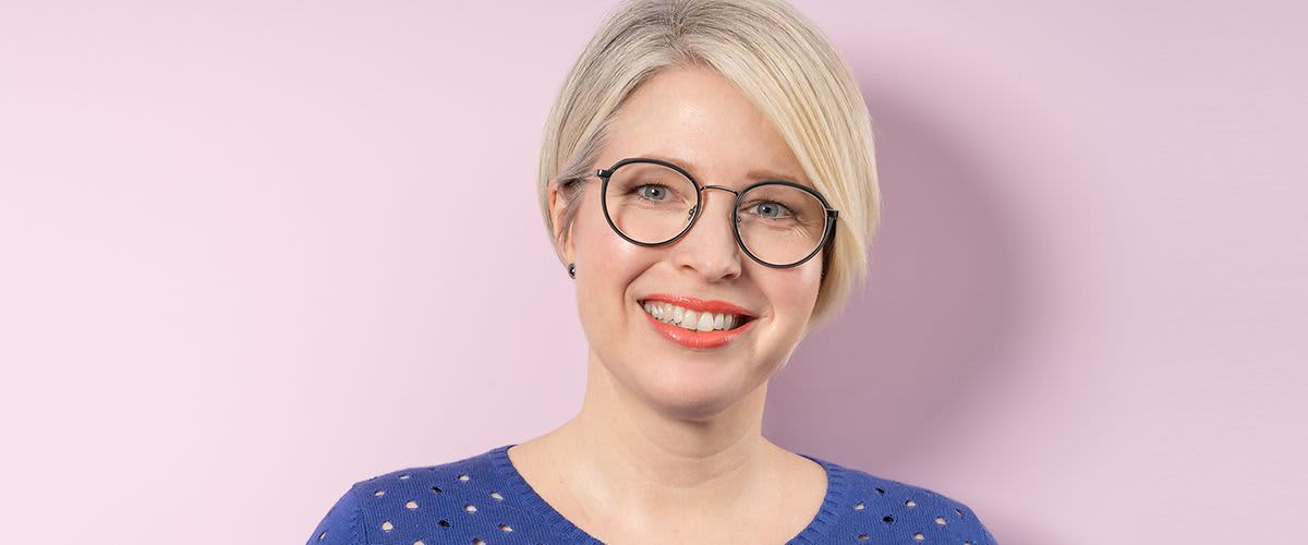 Eine Person mit kurzem, blondem Haar trägt ein blaues Oberteil mit Lochmuster vor einem rosa Hintergrund