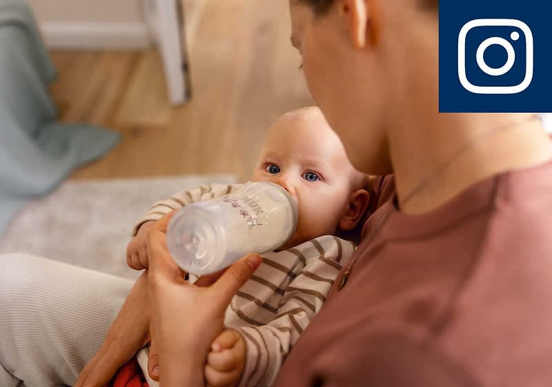 Eine erwachsene Person hält ein schlafendes Baby im Arm und füttert es mit einer Babyflasche, im Vordergrund sind die Hände und die Flasche gut sichtbar, im Hintergrund Teppichboden und Möbelstücke ohne auffällige Details