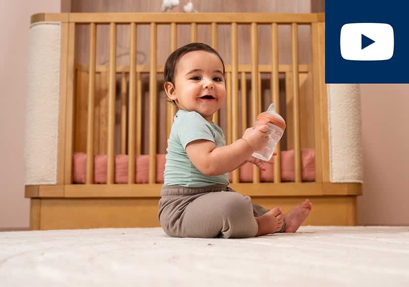 Kleines Kind sitzt auf einem hellen Teppich vor einem Holz-Kinderbett und hält eine Babyflasche in beiden Händen, im Hintergrund weiche Kissen im Bett und rechts oben ein blaues Rechteck mit einem weißen Wiedergabe-Symbol ohne Text