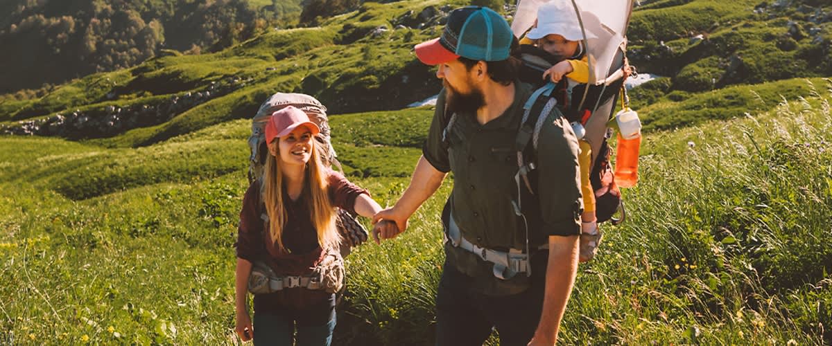 Ein Mann mit Bart und blau-rotem Cap trägt einen Rucksack und hält die Hand eines Kindes, das ebenfalls einen Rucksack trägt, während sie auf einem Wanderweg durch grüne Hügel gehen