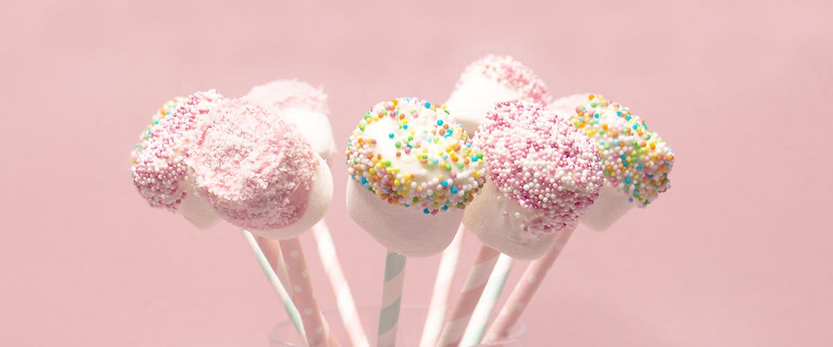 Weiße Marshmallow Pops dekoriert mit bunten Zuckerkugeln auf einem rosanen Hintergrund in einem Glas