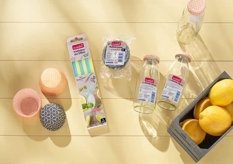Auf einem hellen Holztisch liegen zwei leere Glasflaschen mit rosa gepunkteten Deckeln, eine Packung mit bunten Papierstrohhalmen, zwei Muffinförmchen in orange und schwarz-weißem Muster, ein Paket mit runden schwarzen Muffinförmchen und eine graue Holzkiste mit drei gelben Zitronen.