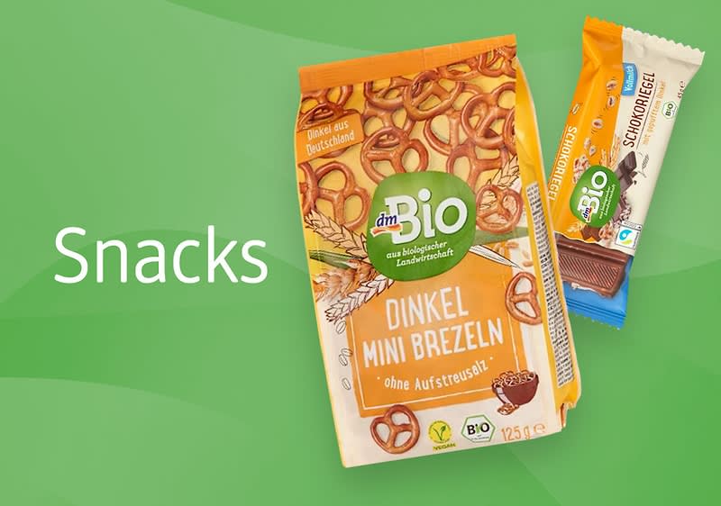 Eine Packung dmBio Dinkel Mini Brezeln mit der Aufschrift „Ohne Aufstreusalz“ und ein dmBio Schokoriegel mit Milchschokolade auf grünem Hintergrund, daneben der Text „Snacks“