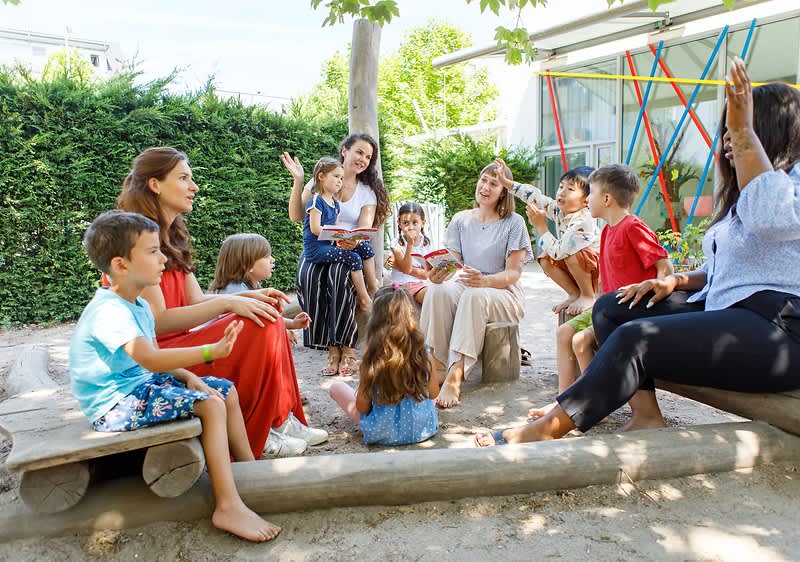 Mehrere Kinder in farbenfroher Kleidung stehen in einem Garten und singen und tanzen gemeinsam mit Erwachsenen