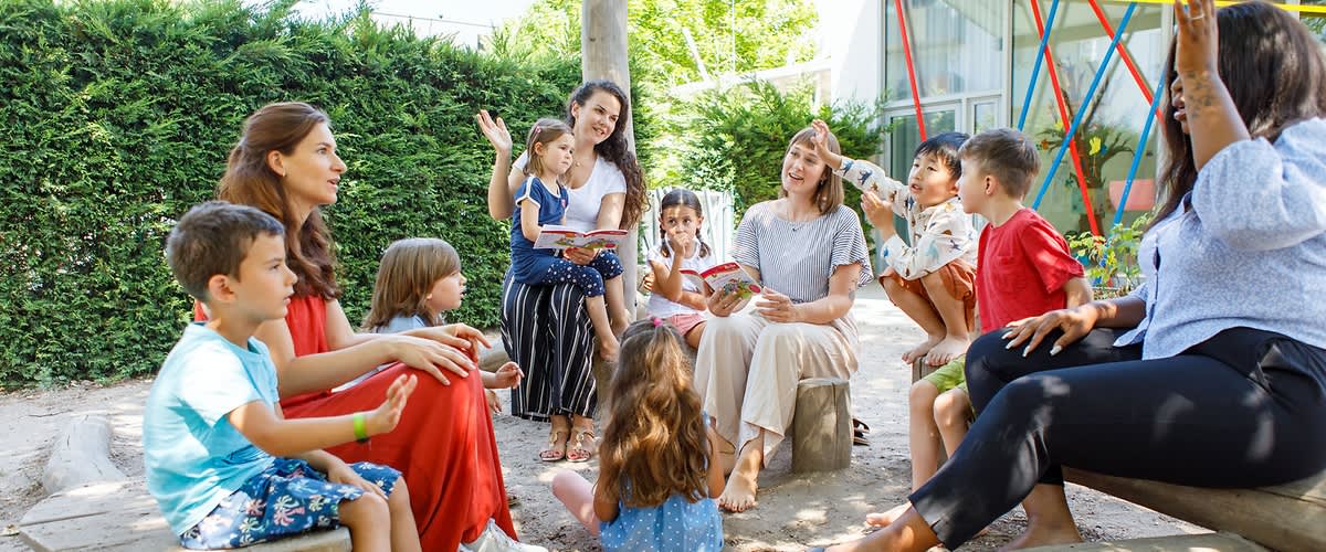 Mehrere Kinder in farbenfroher Kleidung stehen in einem Garten und singen und tanzen gemeinsam mit Erwachsenen