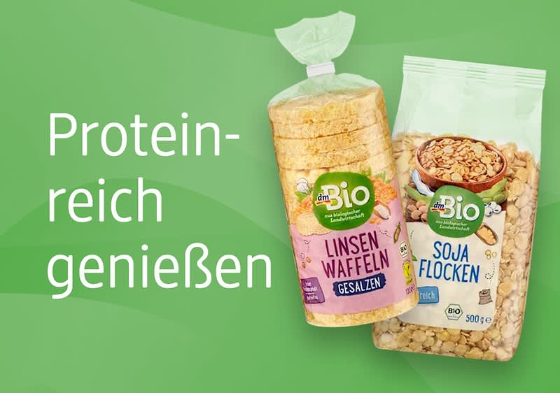 Links steht der Text „Proteinreich genießen“ auf grünem Hintergrund. Rechts sind zwei Produktverpackungen der Marke dmBio zu sehen: Eine Packung Linsenwaffeln mit der Aufschrift „Linsen Waffeln gesalzen“, daneben eine Packung Sojaflocken mit der Aufschrift „Soja Flocken“ und „500 g“