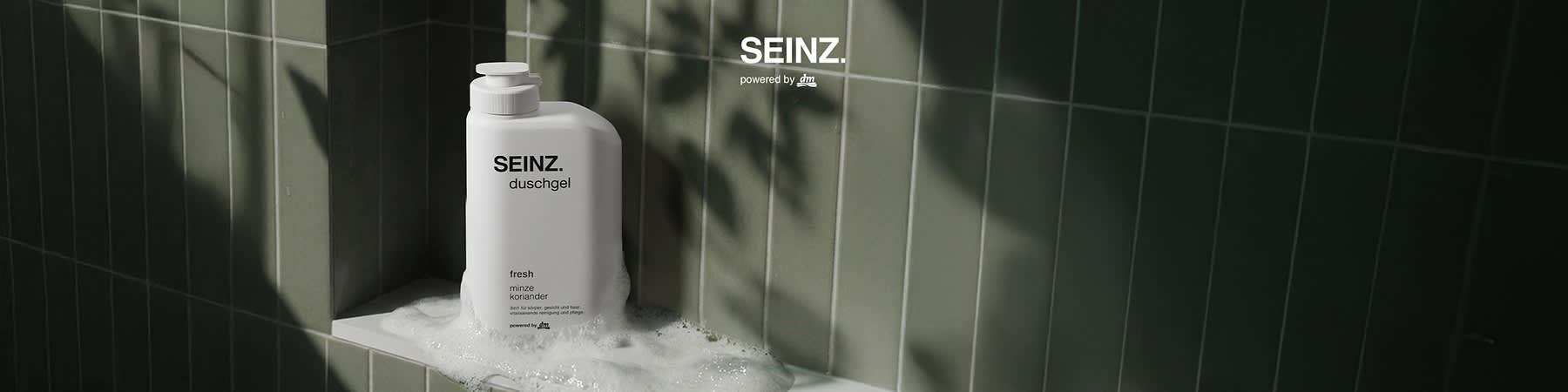 weiße Flasche mit Pumpverschluss der Marke SEINZ. mit Aufdruck „duschgel“ und kleinem Text „fresh mint koriander“, Flasche steht in seifenschaum auf grün gekacheltem Duschbord, oben rechts Schriftzug „SEINZ. powered by dm“. Bild mithilfe von KI erstellt