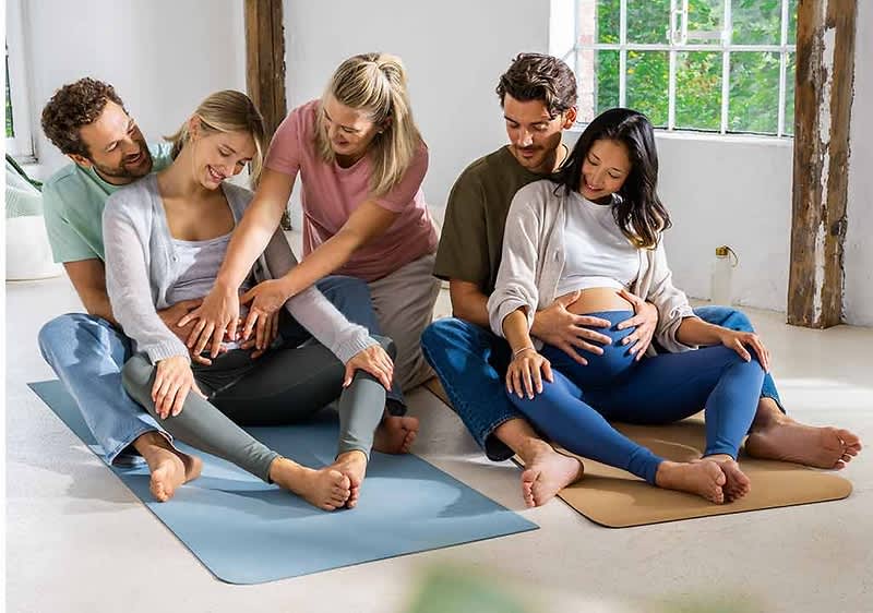 Sechs Personen sitzen in einem hellen Raum auf Yogamatten, zwei schwangere Frauen in der Mitte halten ihre Bäuche, während die anderen vier Personen, zwei Männer und zwei Frauen, unterstützend ihre Hände auf die Schultern und Bäuche der Schwangeren legen, die Fenster im Hintergrund lassen viel Tageslicht herein und die Atmosphäre wirkt entspannt und harmonisch.