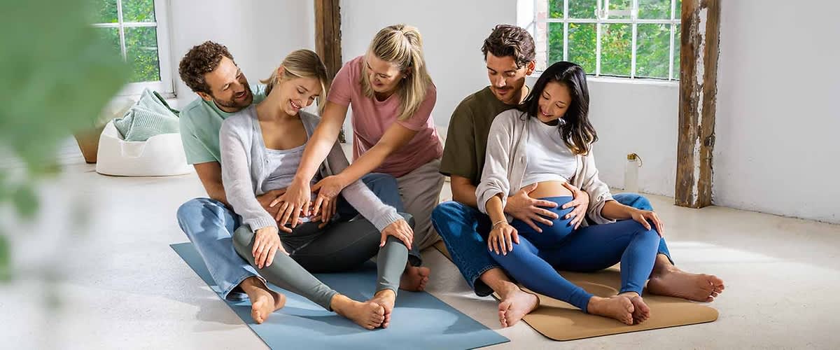 Sechs Personen sitzen in einem hellen Raum auf Yogamatten, zwei schwangere Frauen in der Mitte halten ihre Bäuche, während die anderen vier Personen, zwei Männer und zwei Frauen, unterstützend ihre Hände auf die Schultern und Bäuche der Schwangeren legen, die Fenster im Hintergrund lassen viel Tageslicht herein und die Atmosphäre wirkt entspannt und harmonisch.