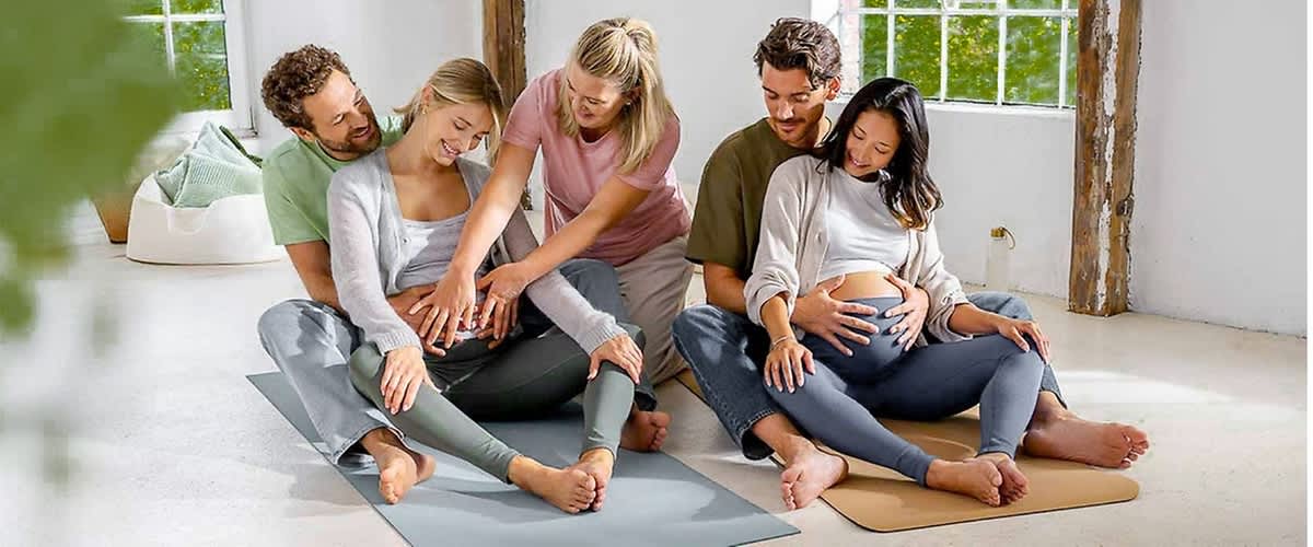 Sechs Personen sitzen in einem hellen Raum auf Yogamatten, zwei schwangere Frauen in der Mitte halten ihre Bäuche, während die anderen vier Personen, zwei Männer und zwei Frauen, unterstützend ihre Hände auf die Schultern und Bäuche der Schwangeren legen, die Fenster im Hintergrund lassen viel Tageslicht herein und die Atmosphäre wirkt entspannt und harmonisch.