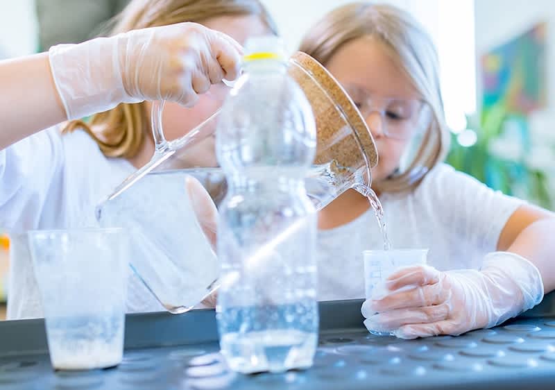 Zwei Kinder mit hellbraunen Haaren und weißen T-Shirts führen ein Experiment durch, indem sie mit transparenten Plastikbechern und einer Flasche arbeiten, während sie weiße Handschuhe tragen; die Flasche ist durchsichtig und enthält eine klare Flüssigkeit, die in die Becher gegossen wird, wobei der Hintergrund unscharf ist und Pflanzen sowie ein Gemälde zu erkennen sind.