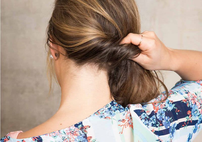 Eine Frau mit blonden Haaren, die im Nacken zu einer Rolle eingeschlagen werden, trägt ein buntes, florales Oberteil und hält die Haare mit einer Hand fest