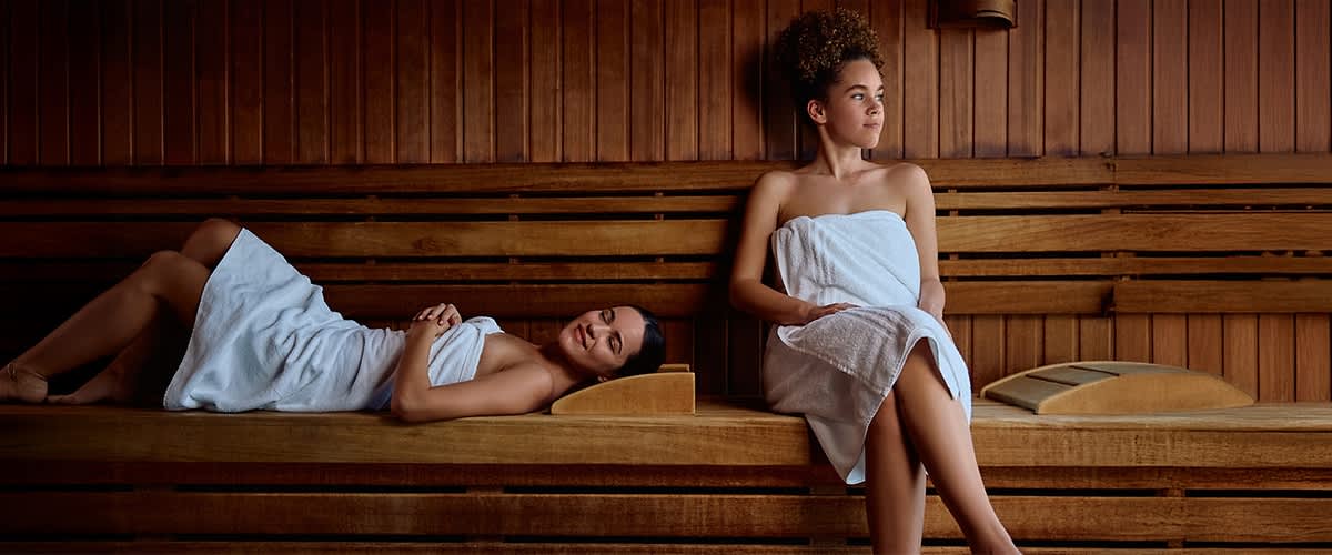 Zwei Personen entspannen in einer Sauna, beide in weiße Handtücher gehüllt. Eine Person sitzt aufrecht, während die andere liegt. Die Sauna ist aus Holz gefertigt.