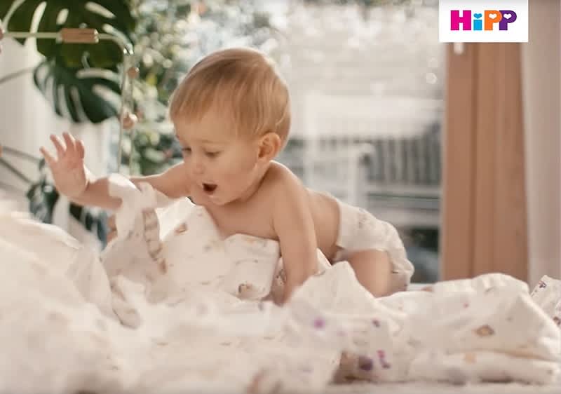 Ein Baby in einer Windel spielt auf einem Bett mit weißer Bettwäsche, im Hintergrund sind Pflanzen und ein Fenster zu sehen, das Logo von HiPP ist oben rechts im Bild