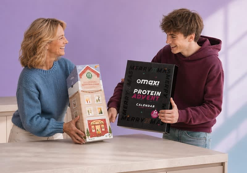 Eine Frau und ein junger Mann sitzen auf einem Sofa und lächeln sich an; die Frau hält einen sechseckigen Bio-Gewürz-Adventskalender in Hausform, der junge Mann hält einen schwarzen Maxi Protein Advent Calendar-Kalender mit pinker Schrift. Bild mithilfe von KI erstellt