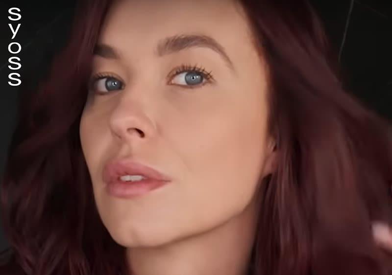 Eine Person mit langen, dunkelroten Haaren, die leicht gewellt sind, vor einem dunklen Hintergrund, wobei das Gesicht unscharf gemacht ist, und links oben steht der weiße Schriftzug Syoss in vertikaler Ausrichtung.