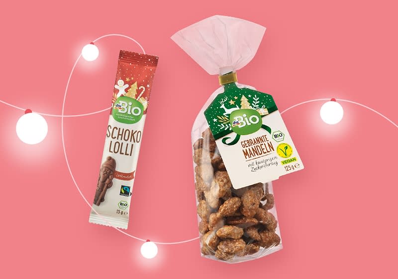 Eine Verpackung mit der Aufschrift „Bio Schoko Lolli“ neben einer Tüte „Bio Gebrannte Mandeln“ auf rosa Hintergrund mit weißen Lichterketten