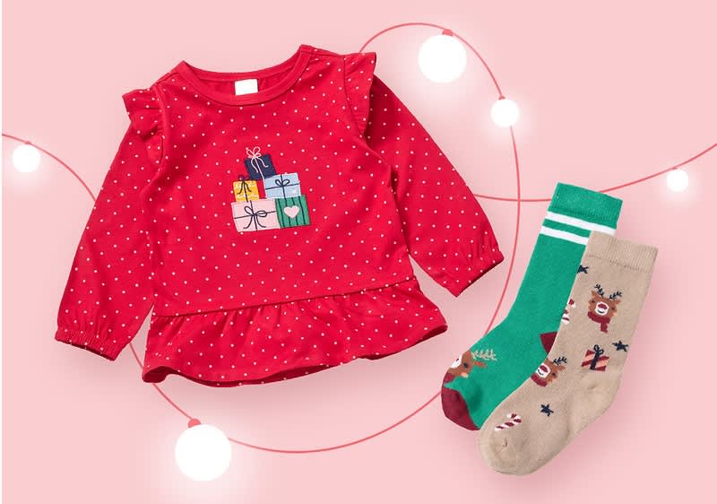 Ein rotes Kinderoberteil mit weißen Punkten und einem Motiv aus bunten Geschenken, daneben zwei Paar Socken, eines in Grün mit Rentiermotiv und das andere in Beige mit Rentier- und Zuckerstangenmotiv