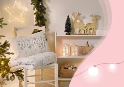 Ein weißer Stuhl mit einem Kissen und einer Decke mit Schneeflockenmuster steht neben einem Regal, das mit zwei goldenen Hirschfiguren, einem kleinen Tannenbaum und zwei beleuchteten Häuschen dekoriert ist. Links sind ein Weihnachtsbaum und eine leuchtende Sternenlichterkette zu sehen. Rechts ist ein rosa Hintergrund mit geschwungenem Rand