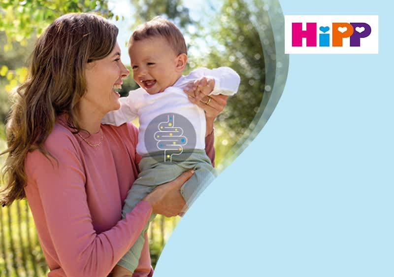 Eine Frau in einem rosa Pullover hält ein Kleinkind in einem weißen Oberteil und grüner Hose im Arm, das Logo von HiPP ist oben rechts im Bild