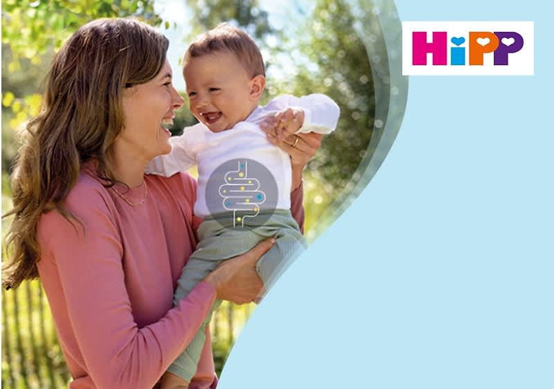 Eine Frau in einem rosa Pullover hält ein Baby in einem weißen Oberteil und grüner Hose, oben rechts das Logo von HiPP