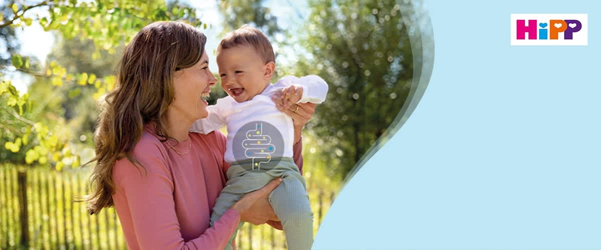 Eine Frau in einem rosa Pullover hält ein Baby in einem weißen Oberteil und grüner Hose, oben rechts das Logo von HiPP