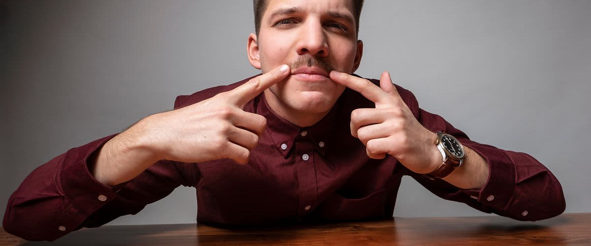Dunkelhaariger Mann mit weinrotem Hemd zeigt mit beiden Fingern auf seinen Schnurrbart.