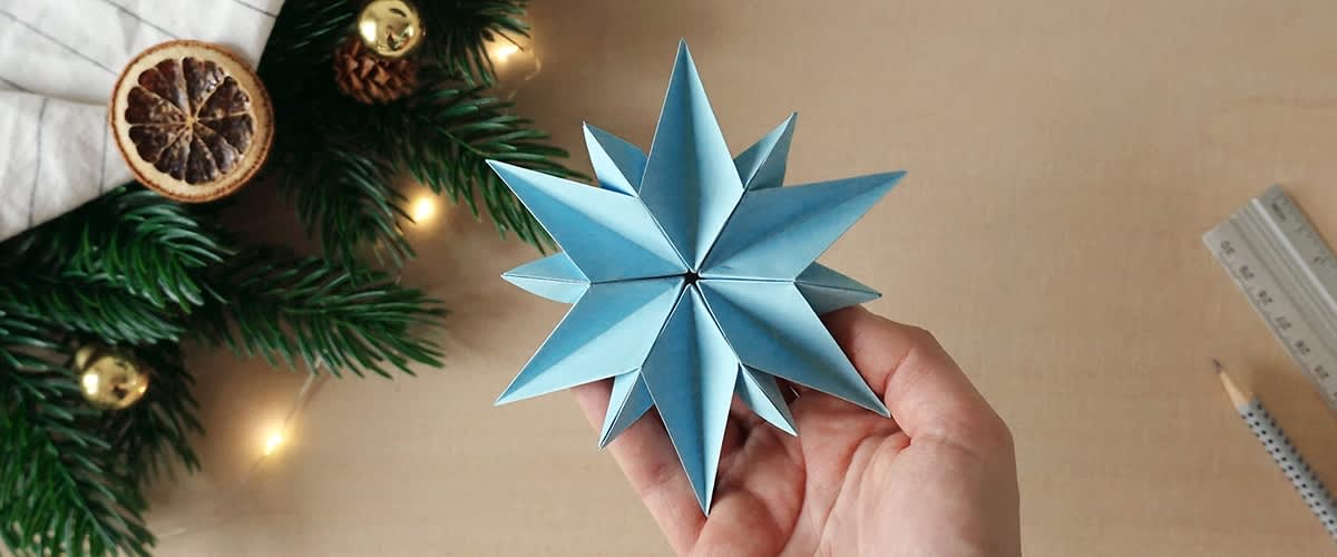 Ruka drži plavu origami zvijezdu, u pozadini su vidljive grane jele s malim lampicama