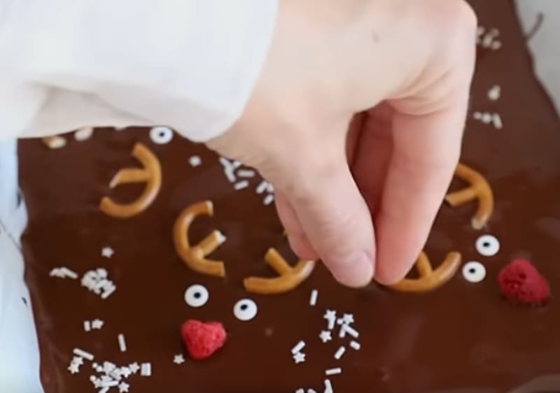 Ruka ukrašava površinu prekrivenu čokoladom, perecima, slatkim očima, malinama i bijelim mrvicama kako bi stvorila lice soba