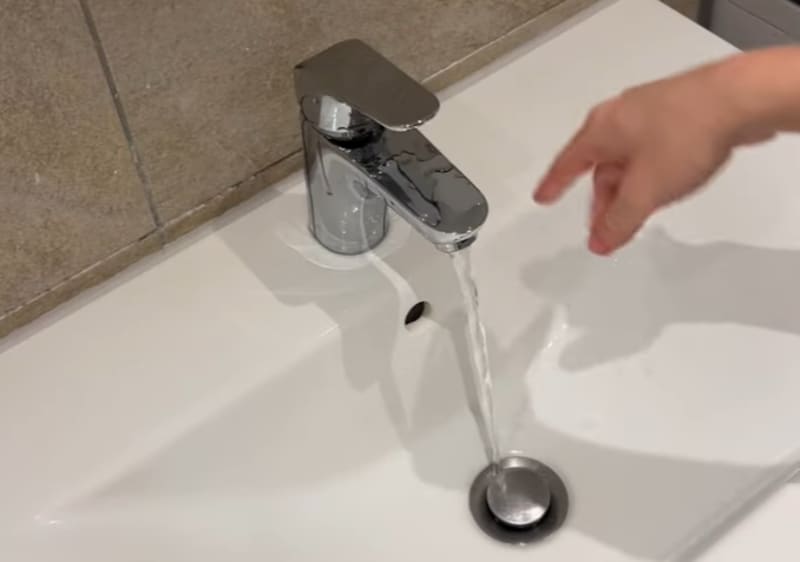 Voda teče iz moderne slavine u bijeli sudoper; pokraj nje se vidi ruka.