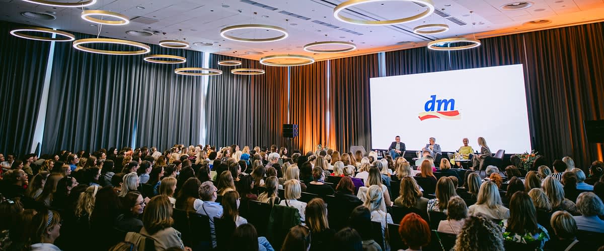 Slika koja prikazuje konferenciju s publikom i panelom govornika ispred ekrana s logotipom dm