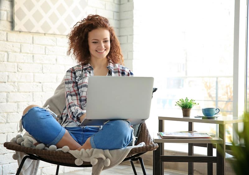 žena koja sjedi kod kuće u fotelji i radi na prijenosnom računalu