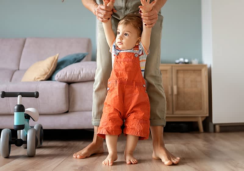 Gyerek narancssárga kantáros nadrágban, felnőtt segíti járás közben, háttérben kanapé és játék tricikli