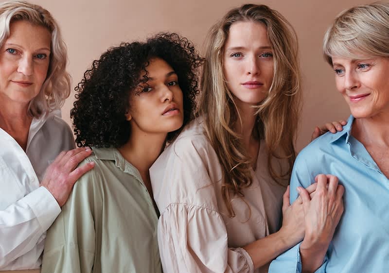Négy különböző bőrszínű nő áll egymás mellett, világos ruhát viselnek, és gyengéden megérintik egymás vállát és karját.