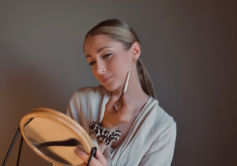 Egy szőke fiatal nő sminkeli magát egy kerek kézitükör előtt