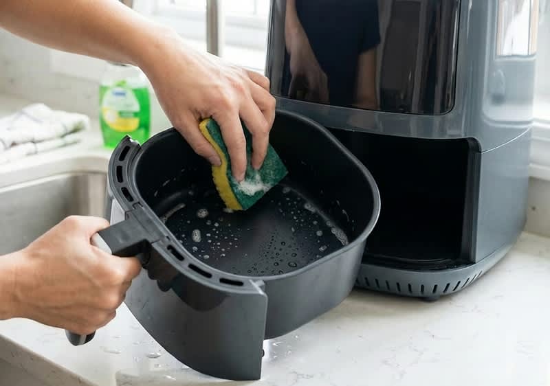 Egy személy szivaccsal és mosogatóvízzel tisztítja a forró levegős fritőz kivett kosarát.