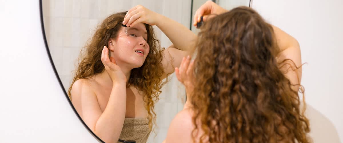Egy fürdőszobatükör előtt álló, vállig érő göndör hajú nő törölközőbe csavarva, az egyik kezével felemelt szérum- vagy olajcseppet visz fel az arcára, miközben a tükörben mosolyogva nézi magát