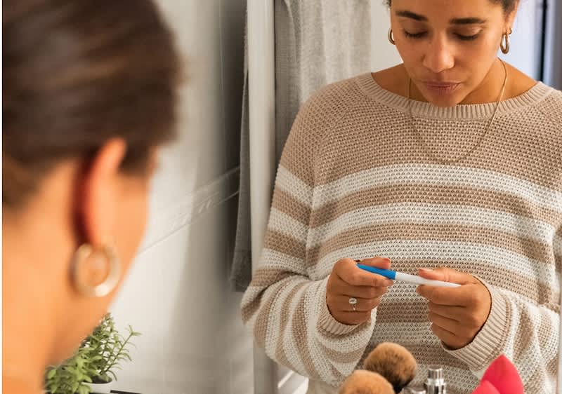 Egy fürdőszobai tükör előtt álló nő közelről, középen egy terhességi tesztet tart két kézzel és nézi, balra a tükörben látszik a feje hátulról, előtérben sminkecsetek és pipetta jellegű eszközök a mosdónál