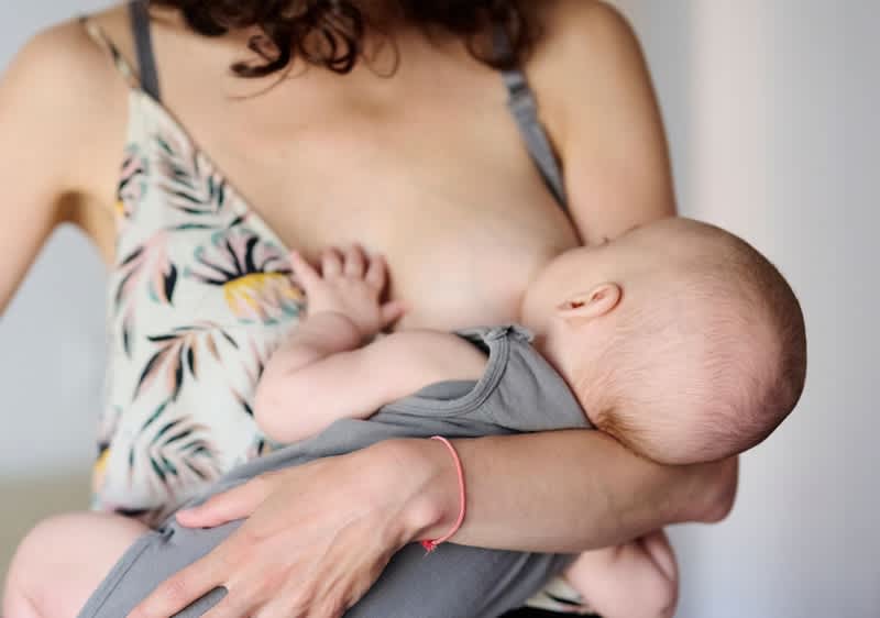Egy ülő anya féloldalasan szoptat egy kisbabát, az anyuka virágmintás felsőt visel és a baba szürke bodyban van, az anya egyik karja a baba fejét és hátát támasztja, a másik kéz a baba lábán pihen, az arcvonások nem jól láthatók