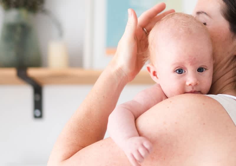 Egy kisbaba fekszik egy nő vállán, a baba kék szemmel előre néz és a nő hátát simítja a keze, a nő sötétbarna hajjal felemelt állásban tartja a gyermeket, ruházatként világos pántú felső látszik a vállon