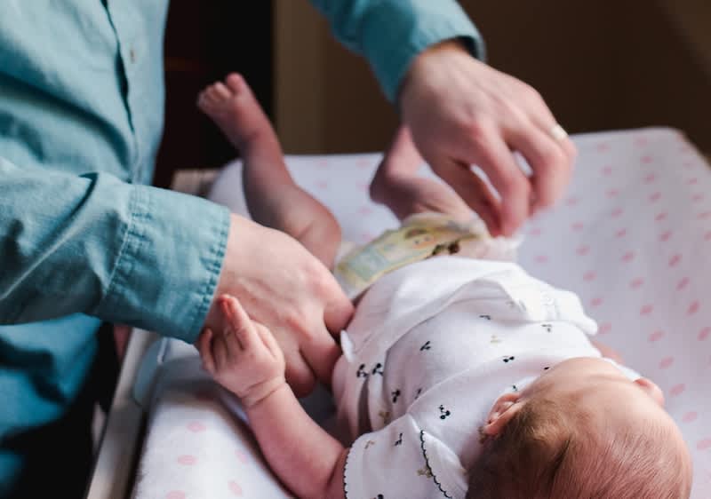 Babát pelenkáz egy felnőtt kézzel; a csecsemő fehér mintás bodyban fekszik pelenkázó lapján, a felnőtt kék ingujjú karja a pelenka rögzítésén dolgozik