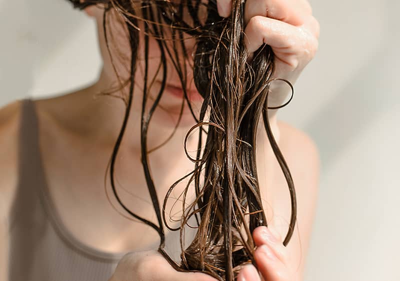 Nedves, sötét hajtincset két kéz fog meg és simít lefelé, a háttérben vállat és részben takart arcot viselő személy látható, világos ujjú topban