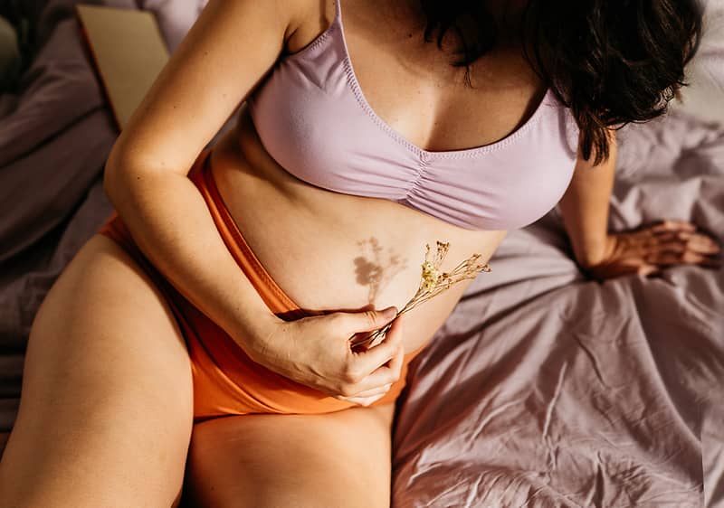 Egy nő ül az ágyon fehér-lilás melltartóban és narancssárga alsóneműben, jobb kezében apró szárított virágot tart az alhas közelében, bal keze az ágyneműn támaszkodik, látható a törzs és combok egy része