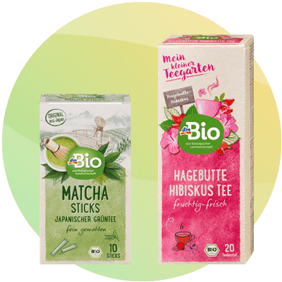 Due confezioni di tè dmBio affiancate su sfondo chiaro: a sinistra confezione verde di Matcha Sticks con scritta Matcha Sticks Japanischer Grüntee e logo dmBio, 10 sticks illustrati; a destra confezione rosa di Hagebutte Hibiskus Tee con logo dmBio, testo fruchtig-frisch e 20 teebeutel