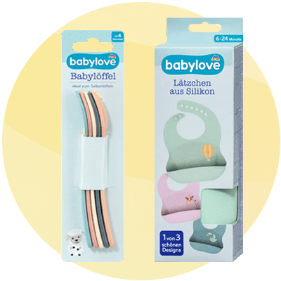Due confezioni del marchio babylove: a sinistra tre cucchiaini flessibili multicolore (rosa, beige, verde) impacchettati su cartoncino azzurro con piccolo disegno pecora; a destra confezione rettangolare di bavaglini in silicone verde e rosa con finestrella e scritta Lätzchen aus Silikon e indicazione età 6–24 Monate