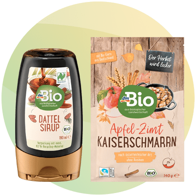 Due confezioni di prodotti dmBio su sfondo chiaro: a sinistra flacone scuro con etichetta beige e scritta Dattel Sirup 180 ml raffigurante datteri; a destra sacchetto beige con grafica legno e frutta e scritta Apfel-Zimt Kaiserschmarrn 140 g e logo dmBio
