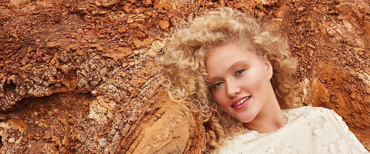 Donna con capelli ricci biondi appoggiata su una superficie rocciosa.
