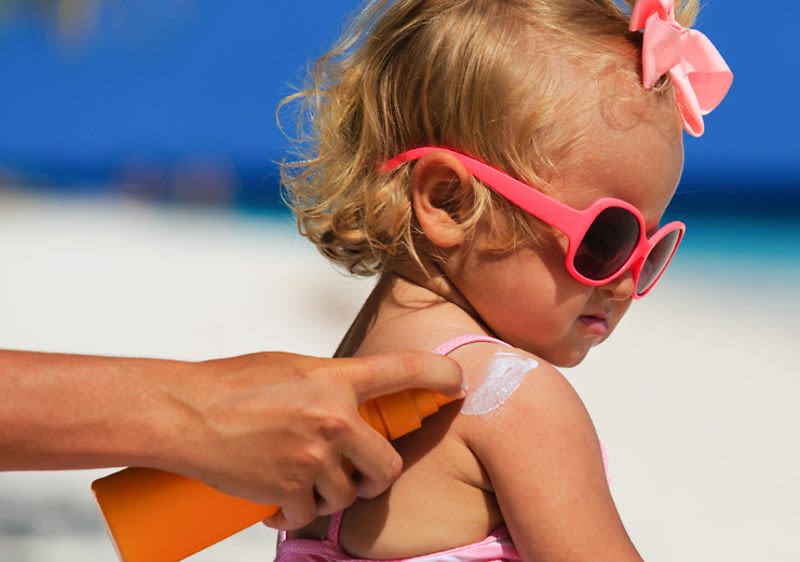Kleines Mädchen mit Sonnenbrille, das eingecremt wird, am Strand